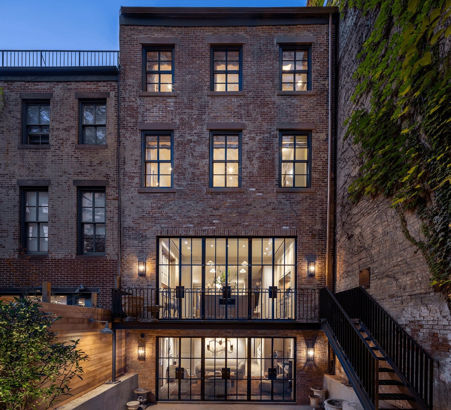 A brownstone rear facade with tall black steel windows and doors, lit at dusk.
