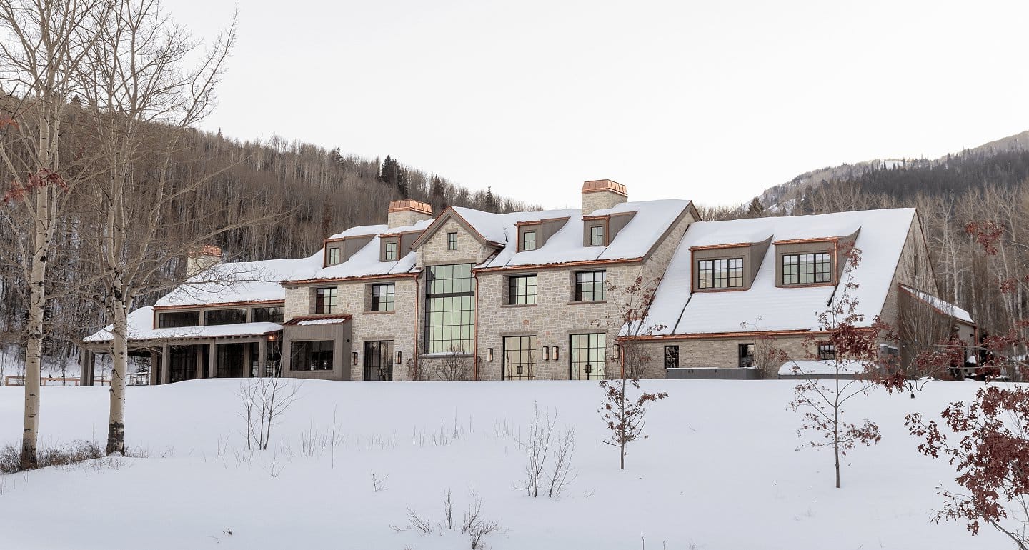 A stone mountain home in snow, its tall steel windows glowing.