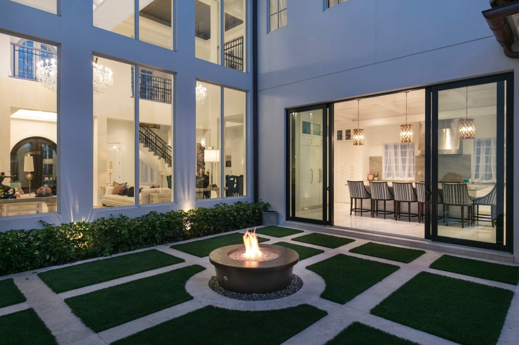 Private courtyard with a lit fire feature, framed by black-framed floor-to-ceiling glazing and a multi-panel sliding glass door opening from the kitchen and living areas.