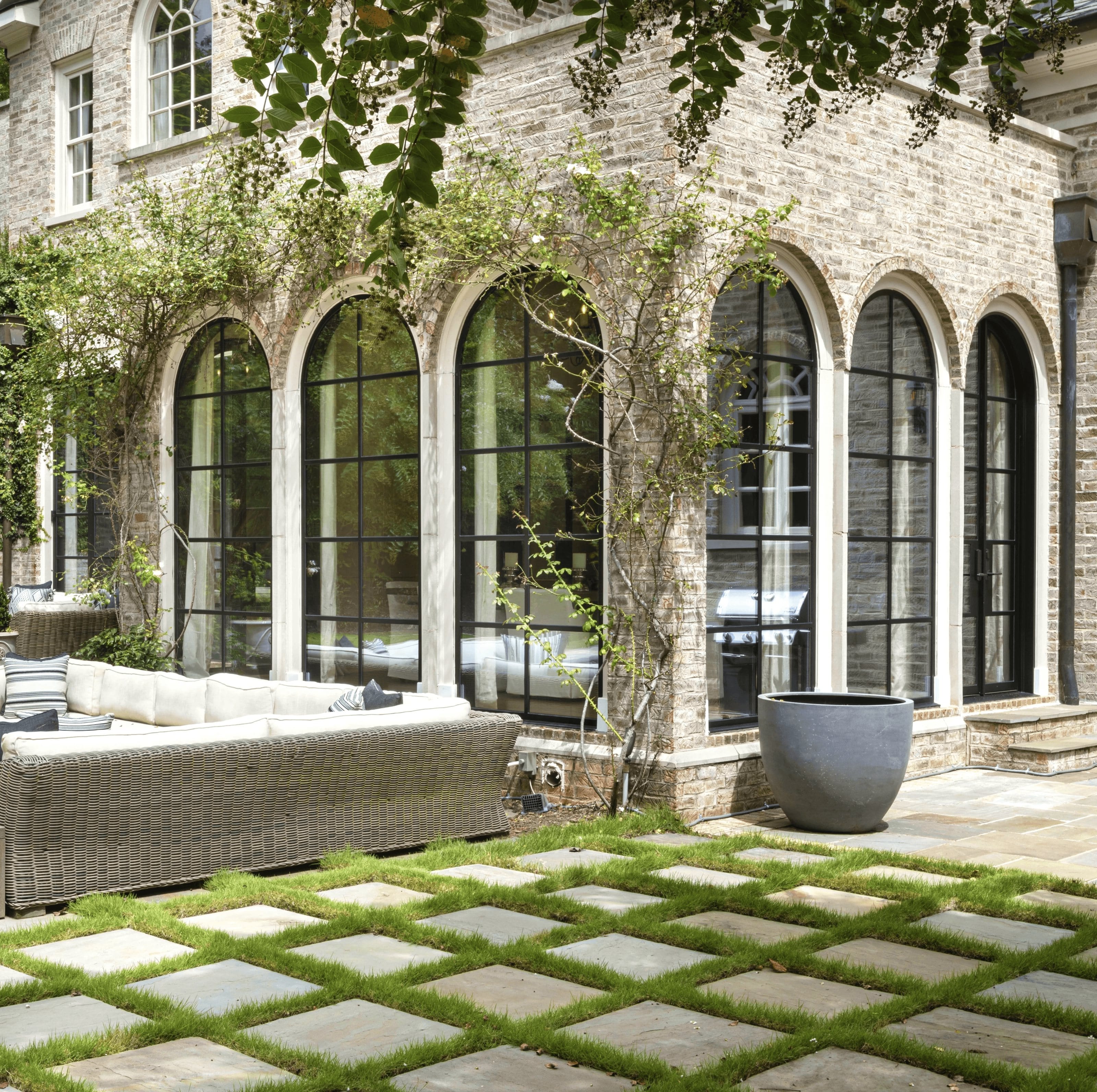 A run of tall arched steel-framed windows set into a limestone facade, seen across a paved courtyard planted with grass joints.